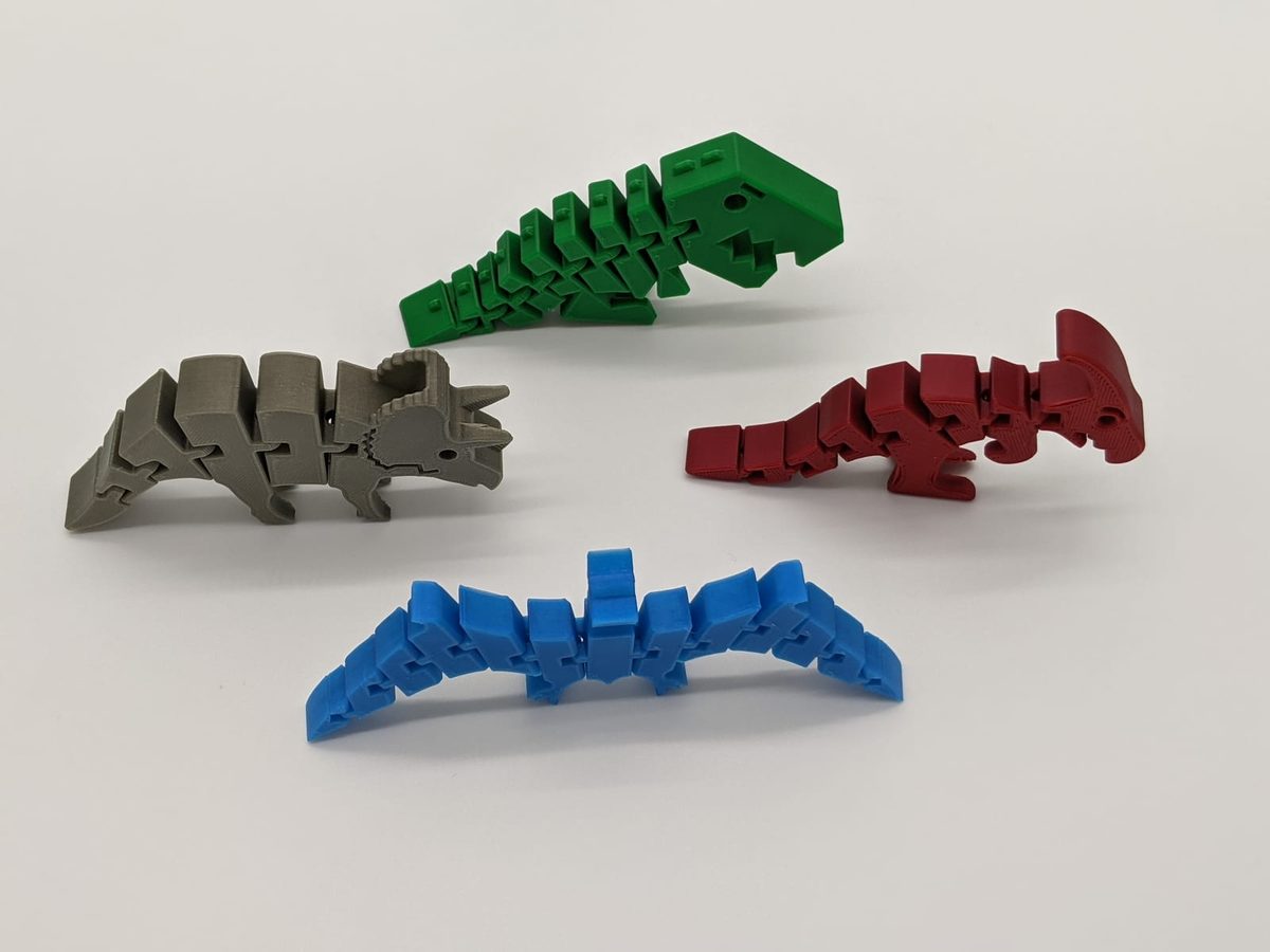Four articulated 3D-printed dinosaurs — a green T-rex, gray triceratops, red raptor and blue stegosaurus